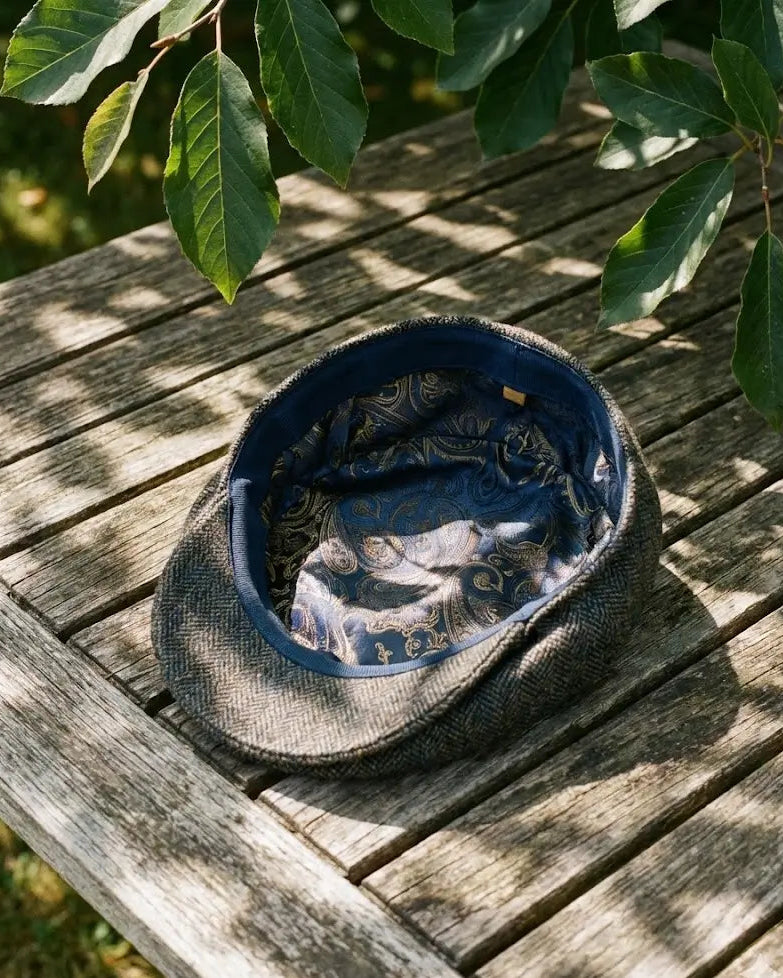 Interior da boina inglesa Cappel exibido sobre uma mesa de madeira. Destaque para o forro de cetim com estampa paisley azul, projetado para ser respirável e oferecer conforto térmico em dias quentes. Acabamento interno de luxo.