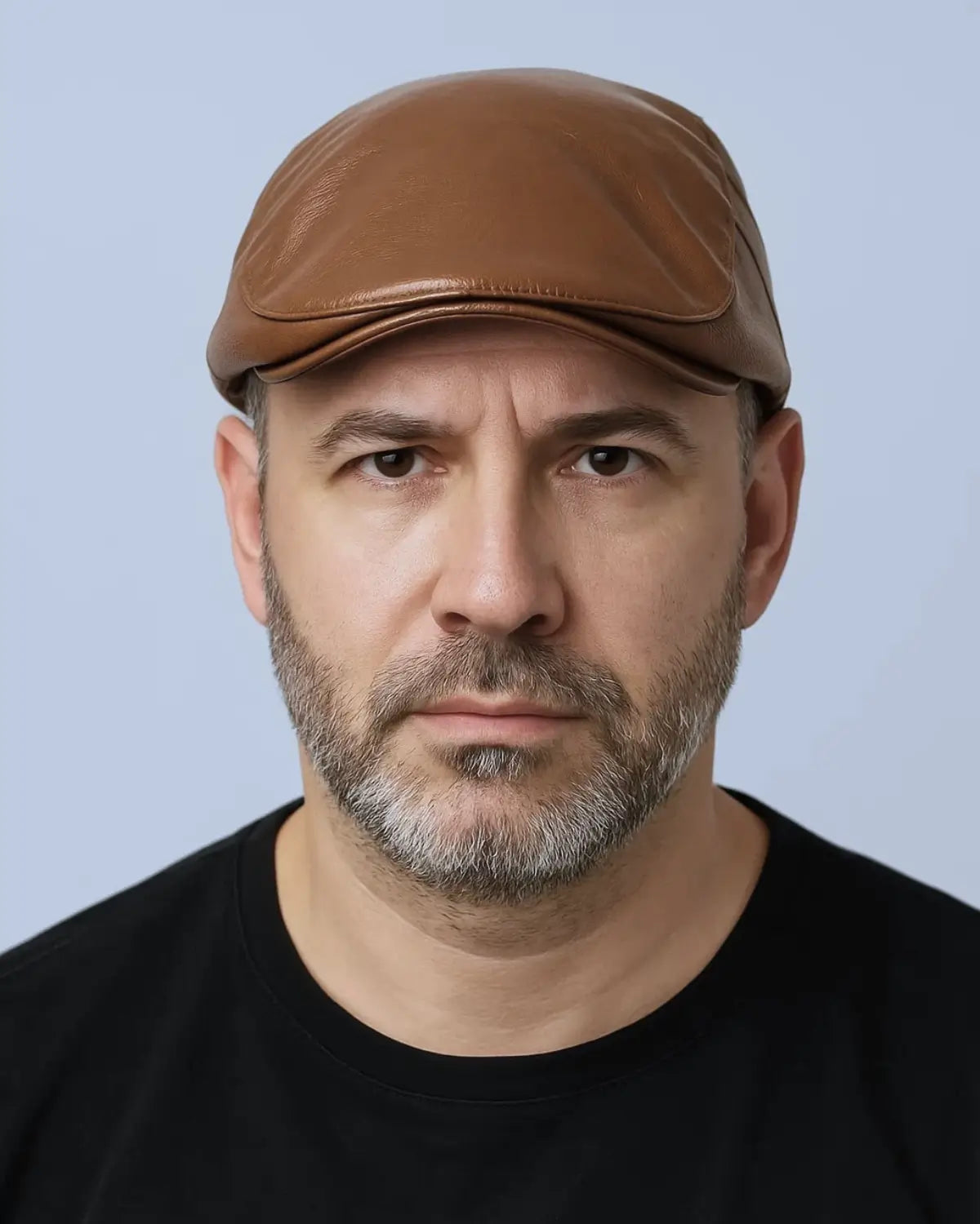 Retrato frontal de homem com boina inglesa marrom e expressão séria, fundo neutro claro