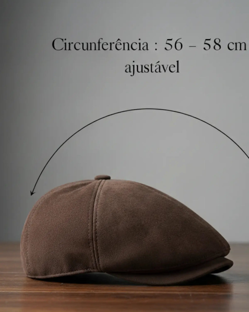 Boina masculina inglesa em couro vegano marrom com indicação de circunferência ajustável de 56 a 58 cm sobre mesa de madeira, acessório sofisticado