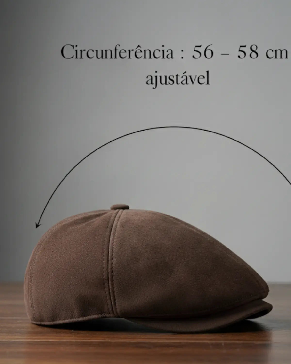 Boina masculina inglesa em couro vegano marrom com indicação de circunferência ajustável de 56 a 58 cm sobre mesa de madeira, acessório sofisticado